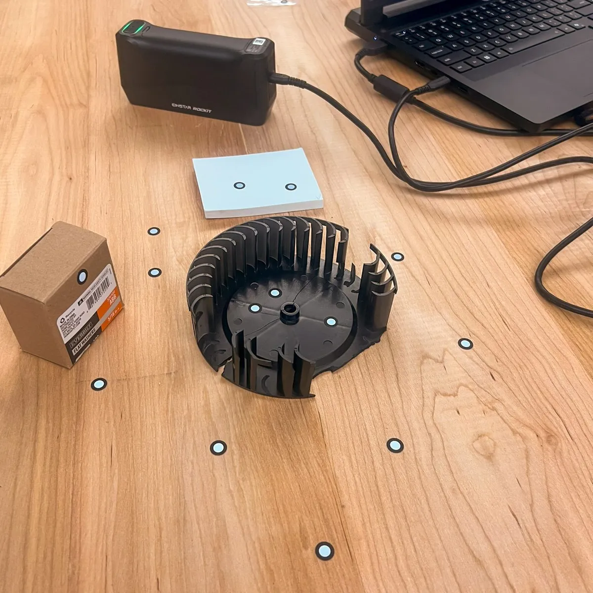 Broken fan wheel on a workbench surrounded by scan reference markers, with a sticky note pad, a box of nails, and a handheld 3D scanner connected to a laptop