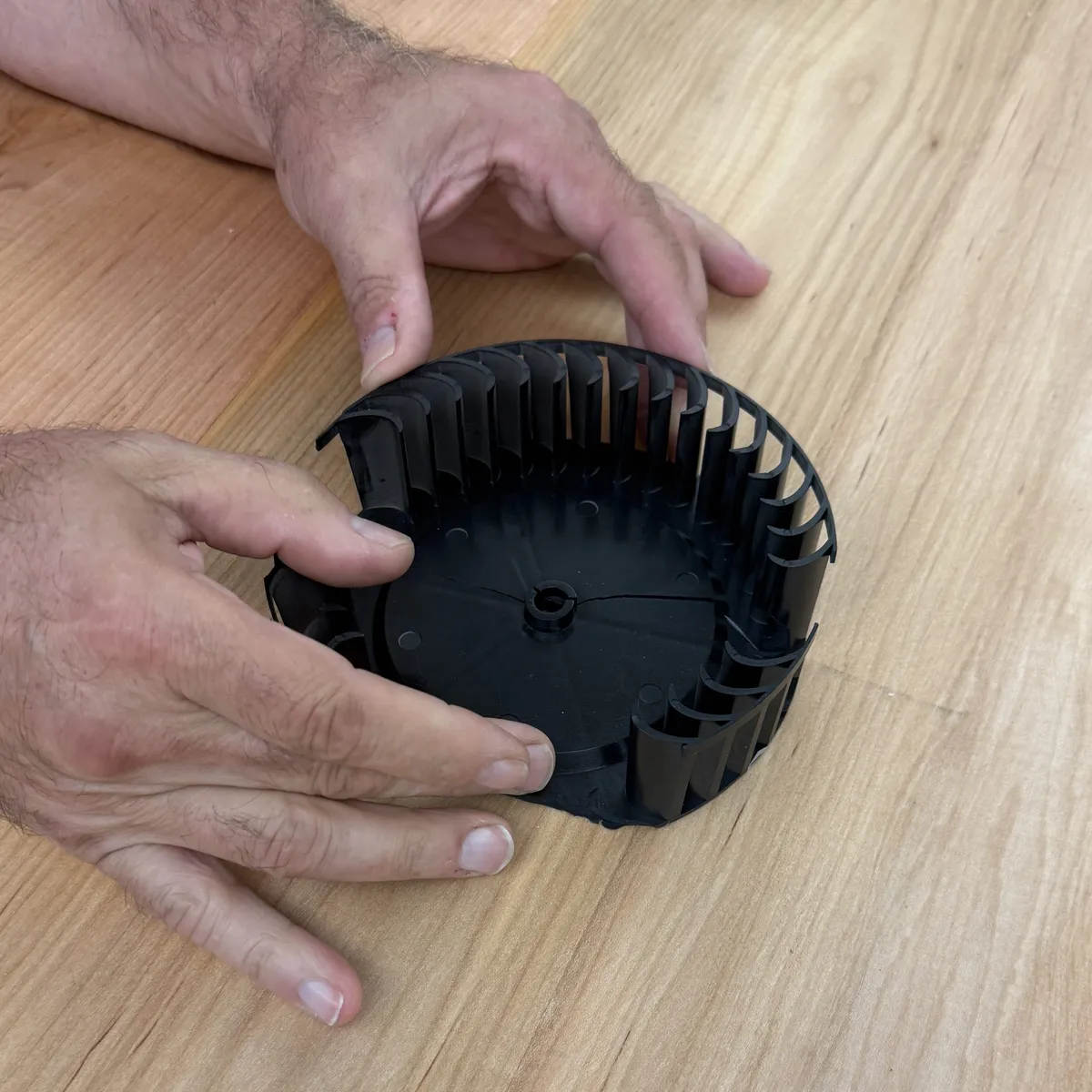 Hands holding the broken black blower fan wheel, with a section of its curved blades missing