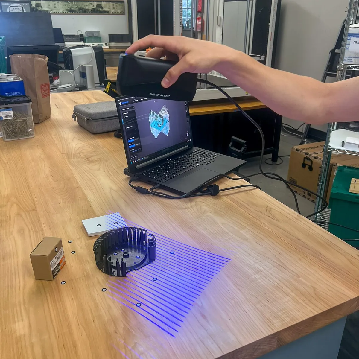 Handheld 3D scanner projecting blue laser lines over the fan wheel while the live scan appears on the laptop screen